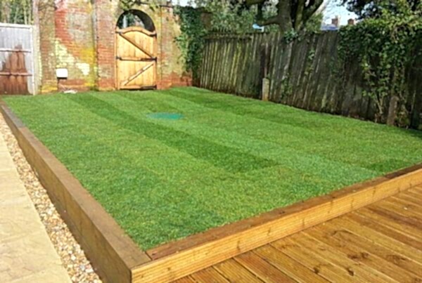 A freshly mowed, manicured turf is bordered by a wooden fence and an old brick wall with a wooden gate. The lawn is elevated with wooden planks, separated from a wooden decked area. Trees and greenery overhang the shaded corner of the yard.