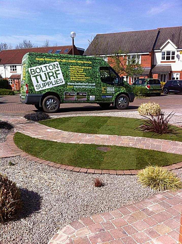 A Bolton Turf Supplies van, adorned with lush turf graphics and company contact details, is parked on a landscaped residential street with houses and gardens visible in the background. The scene includes well-maintained lawns, vibrant plants, and a paved driveway.