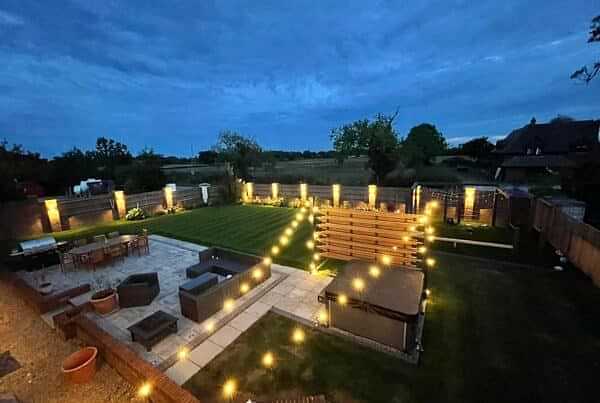 A backyard at dusk adorned with string lights creates a warm atmosphere. The yard features a patio with outdoor furniture, a hot tub, well-manicured turf laid to perfection, and a dining area. The sky is a deepening blue, and trees and fencing frame the spacious yard.