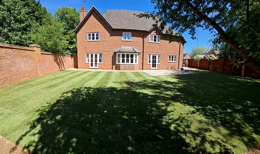 A two-story brick house with a large, neatly mowed turf lawn in the backyard. The yard is surrounded by a brick wall and trees, with a clear blue sky overhead. There is a small patio area to one side of the lawn.