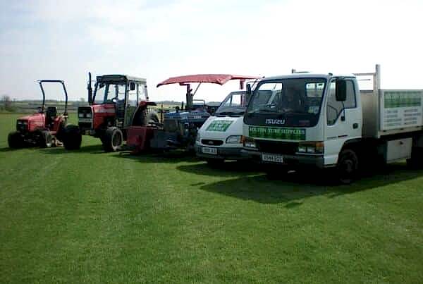 Four vehicles parked on a grassy field. From left to right: a red tractor with a cabin, a tractor with a red canopy, a white van with a raised hood, and a white flatbed truck with green writing on the side—ready for some turf laying. The sky above is clear and blue.